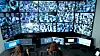 A female security operator wearing a headset monitors multiple surveillance camera feeds on large screens in a control room. She is focused on her workstation, communicating and analyzing real-time data to ensure safety and security. The scene highlights modern security systems, video surveillance technology, monitoring operations, and professional workplace environments in a high-tech setting.