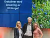 Tre personer står foran en blå skjerm med teksten «Gratulerer med lansering av KI Norge!» og et grønt blomsterbilde.